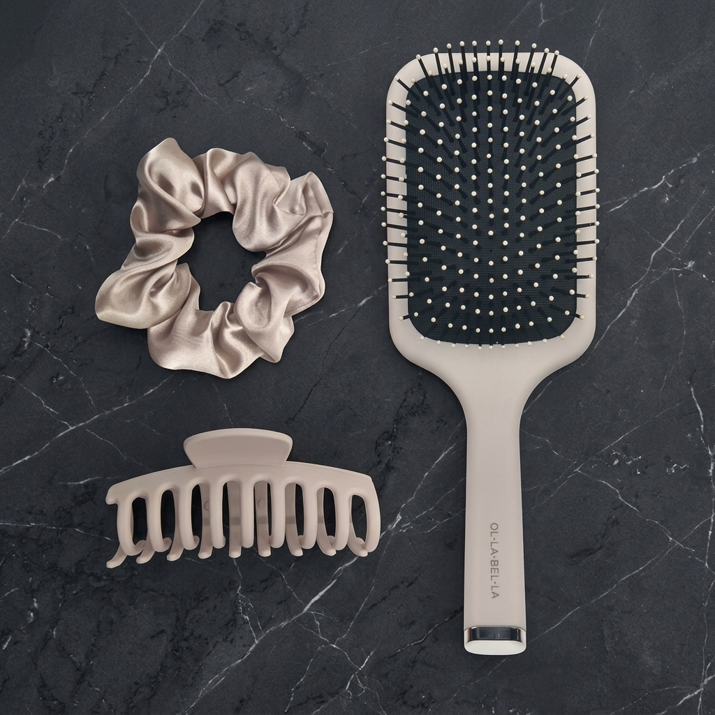 The Olla Bella Everyday Glam Hair Accessory Kit featuring a satin scrunchie, a claw hair clip, and a paddle hairbrush arranged on a dark gray marble background.