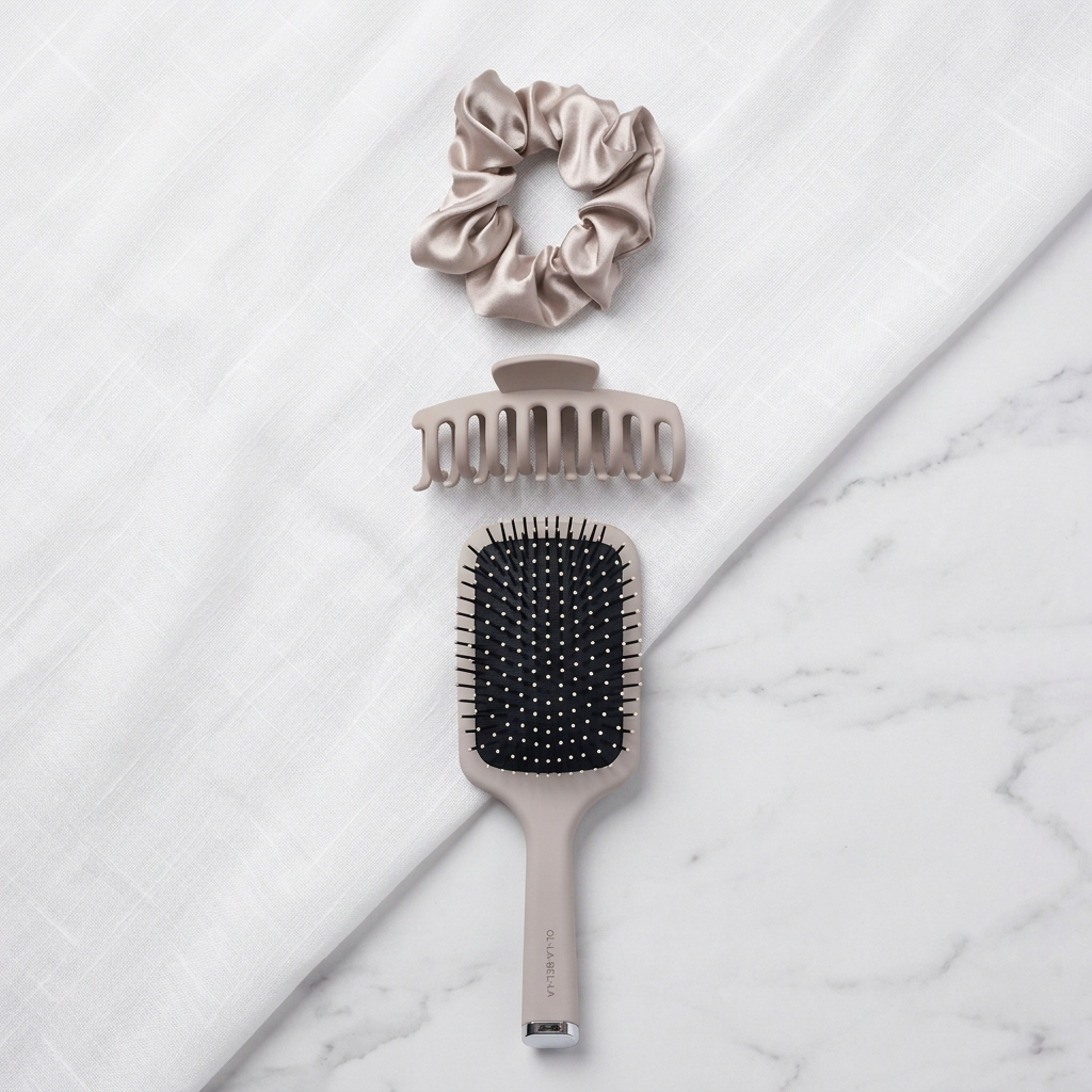 Three haircare accessories, the Olla Bella Everyday Glam Hair Accessory Kit, arranged vertically on a white textured fabric over a white marble surface: a satin scrunchie, a claw comb, and a paddle brush.  The brand name Olla Bella is subtly printed near the base of the brush handle.