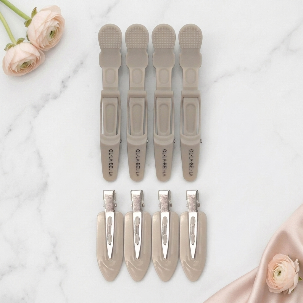 Four Olla Bella Crocodile Clips and four Olla Bella Creaseless Clips from our Olla Bella Mane Master Hair Clip Set arranged in two rows of two on a light marble background with blush-pink flowers and a subtle piece of blush satin fabric.