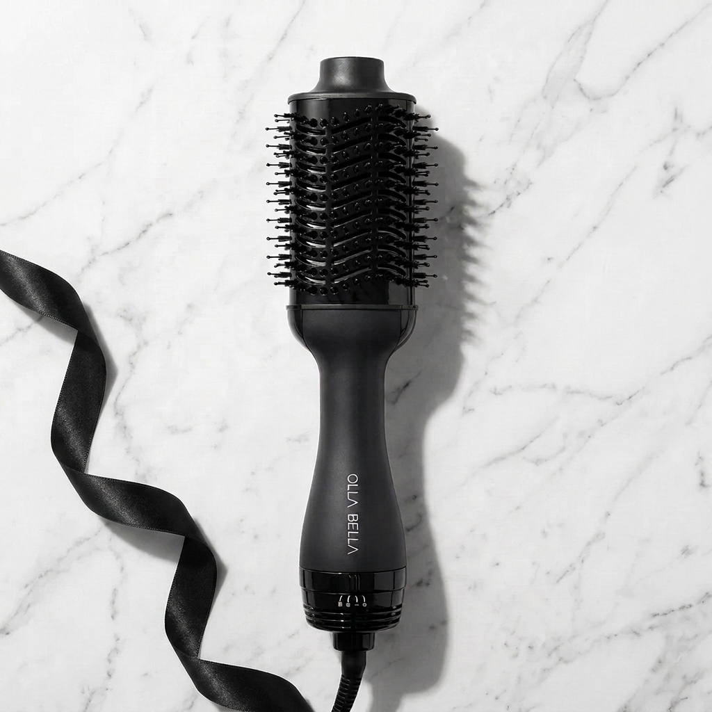 The black Olla Bella Volumizing Blowout Brush with a round barrel and bristle head, shown on a white marble background with a black ribbon accessory.